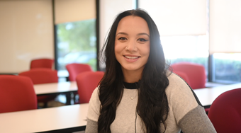 Student Emaree Stone shown in white shirt in a well lit Salus classroom with red chairs, white tables, and windows in the background.